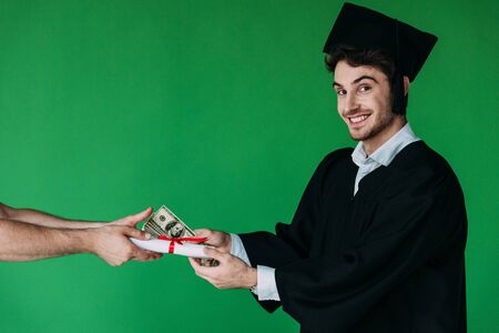 student in academic cap holding diploma with red ribbon and offering bribe isolated on greenの写真素材