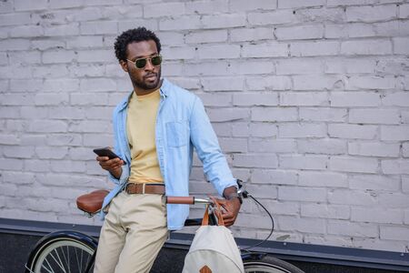 stylish african american businessman in sunglasses standing with bicycle near brick wall and holding smartphoneの写真素材