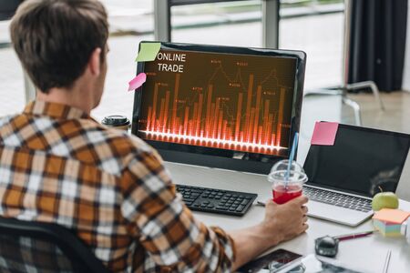back view of young programmer holding glass of juice while sitting at workplace near computer...
