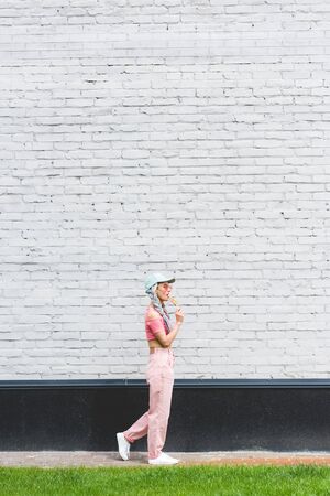 side view of girl in hat eating lollipop near brick wallの写真素材