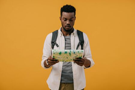 handsome african american man with backpack looking at map isolated on orangeの写真素材