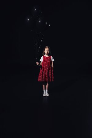 sad child standing with black balloons and looking at camera isolated on blackの写真素材