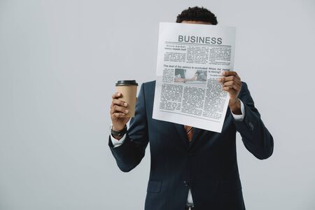 african american man holding disposable cup and covering face while reading business newspaper isolated on greyの写真素材