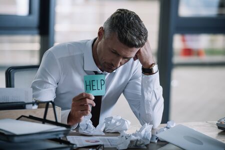 selective focus of upset businessman holding sticky note with help lettering near table