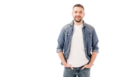 front view of smiling bearded man in denim shirt standing with hands in pockets isolated on white