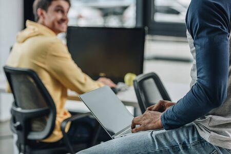 partial view of african american programmer sitting on desk with laptop near smiling colleagueの写真素材