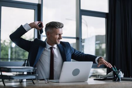 selective focus of upset businessman in suit holding smartphone with blank screen near laptop in officeの写真素材