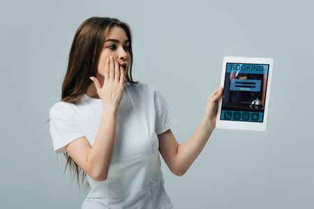 shocked beautiful girl in white t-shirt showing digital tablet with booking app isolated on grey