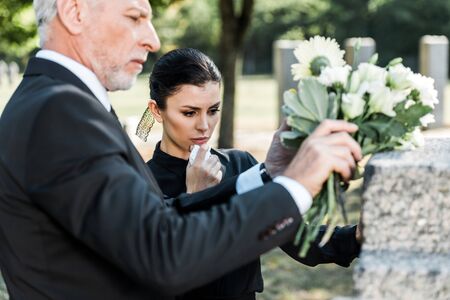 selective focus of senior man holding flowers near tomb and womanの写真素材