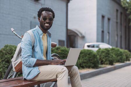 cheerful african american man smiling at camera while sitting on bench and using laptopの写真素材