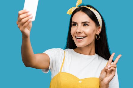 selective focus of happy woman taking selfie on smartphone while showing peace sign isolated on blue