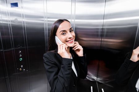 cheerful businesswoman talking on smartphone in elevator and smiling at cameraの写真素材