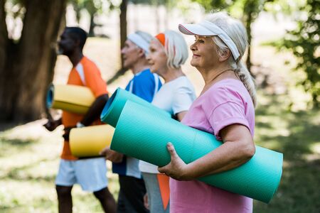 selective focus of happy retired woman holding fitness mat near multicultural pensioners in sportswearの写真素材