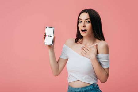 KYIV, UKRAINE - JUNE 6, 2019: beautiful shocked girl holding smartphone with tinder app isolated on pinkのeditorial素材