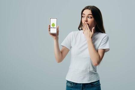 KYIV, UKRAINE - JUNE 6, 2019: shocked beautiful girl in white t-shirt showing smartphone with android logo isolated on greyのeditorial素材