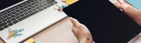 cropped view of woman holding digital tablet in hands over wooden table with laptop, planners and...