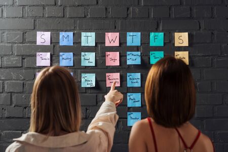 back view of two women standing near colorful sticker notes and pointing with finger on black brick...