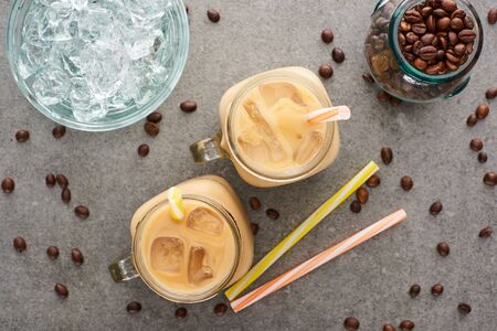 top view of ice coffee in glass jars near straws, coffee grains and ice cubes on grey backgroundの写真素材