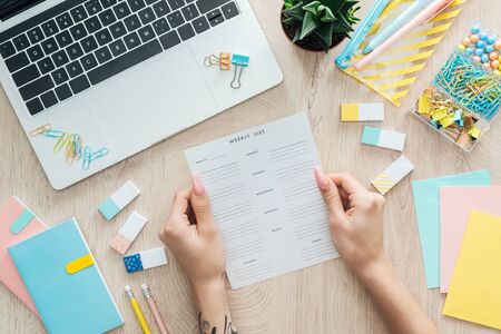 cropped view of woman sitting behind wooden table with laptop and stationery, holding in hands...