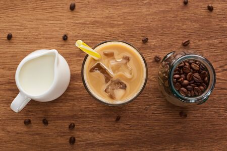 top view of ice coffee with straw near milk in jug and coffee grains in jar on wooden tableの写真素材