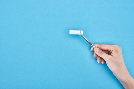cropped view of woman holding tweezers with cotton pad on blue