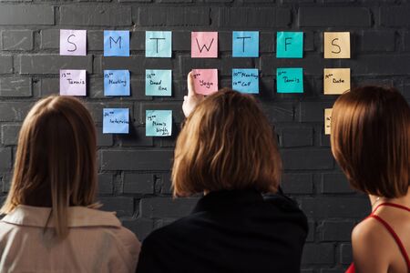 back view of three women standing near colorful sticker notes and pointing at black brick wallの写真素材