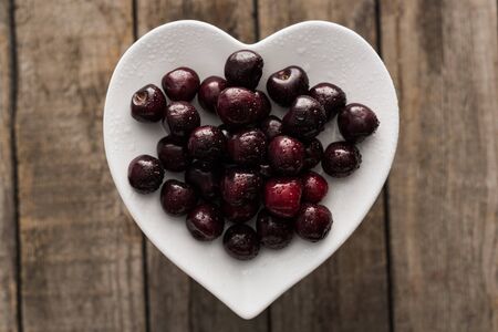 top view of red, fresh, whole and ripe cherries covered with droplets on plate on wooden surface