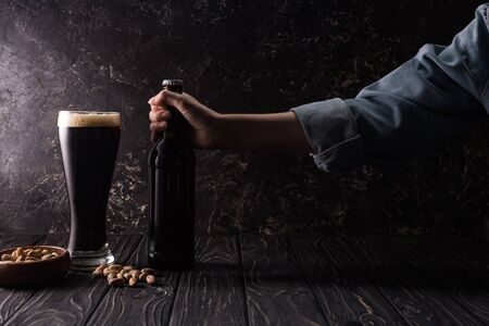 cropped view of man taking bottle near glass of beer and bowl with pistachios on wooden table