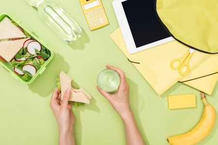 cropped view of woman hands with sandwich and glass of water near lunch box, backpack, digital tablet, bottle of water and stationeryの写真素材