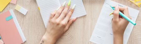 cropped view of woman writing in weekly list, sitting behind wooden table with notepad and...