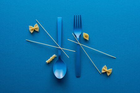 flat lay of spaghetti on blue plastic spoon upside down and fork near different kinds of uncooked pasta on blue backgroundの写真素材