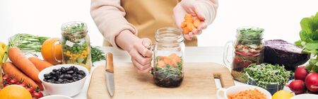 cropped view of woman in apron adding carrots to glass jar with beans on wooden table isolated on white, panoramic shotの写真素材