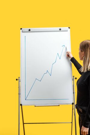 businesswoman standing near white flipchart, pointing at growth graphic, standing isolated on yellowの写真素材