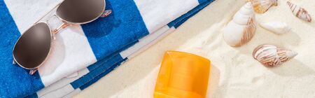 orange bottle of sunscreen on sand with seashells, striped towel and sunglasses, panoramic shot