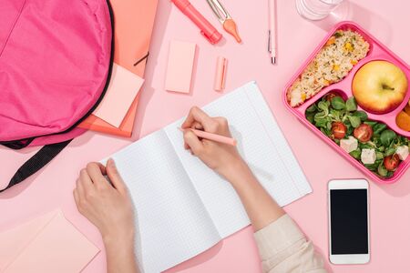 cropped view of woman writing in notebook near backpack and stationery