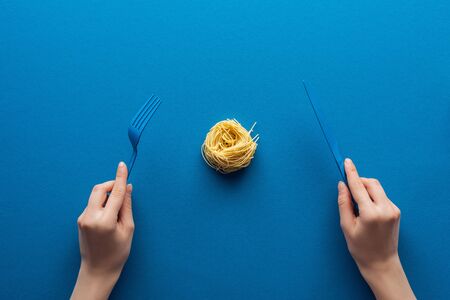 cropped view of woman holding blue plastic fork and knife with vermicelli pasta in middle on blue backgroundの写真素材