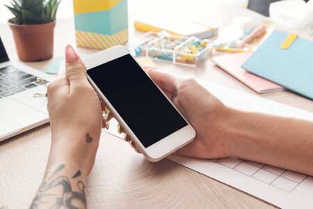 cropped view of woman sitting behind wooden table with laptop and stationery, holding smartphone in...