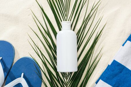 white sunscreen lotion on green palm leaf on sand with blue flip flops and towelの写真素材