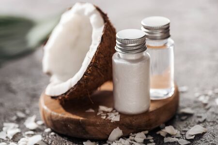 selective focus of bottles with coconut beauty products on wooden board near coconut half on grey...