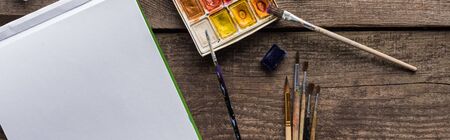 top view of colorful paint palette, paintbrushes and blank sketch pad on wooden surface, panoramic shotの写真素材