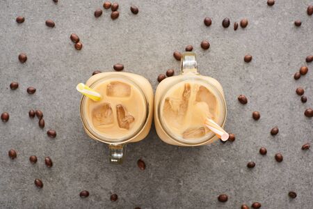 top view of ice coffee in glass jars with straws and coffee grains on grey background
