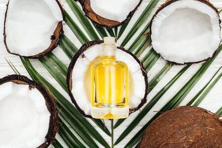 top view of coconut oil in bottle on coconut half and green palm leaf on white wooden surfaceの写真素材