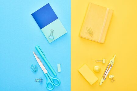 Top view of yellow and blue notepads with different stationery on bicolor background