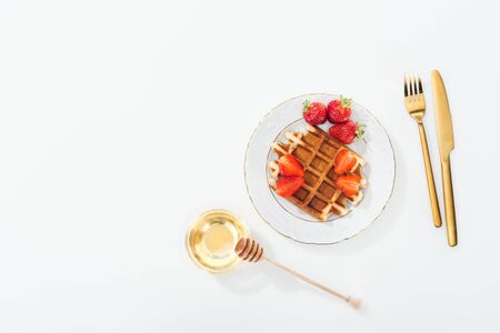 top view of waffle with strawberries on plate near bowl with honey and wooden dipper, and cutlery on whiteの写真素材