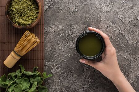 top view of woman holding matcha matcha tea on table with whisk, powder and mint