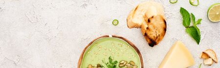 panoramic shot of tasty green creamy soup with croutons and cheese on textured grey background