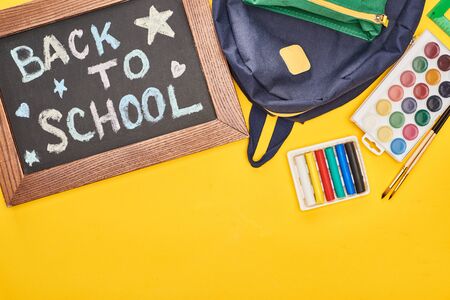 Chalkboard in wooden frame with back to school inscription near blue schoolbag and watercolor paints...