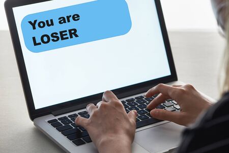 cropped view of girl typing on laptop keyboard with you are loser message on screen, cyberbullying...