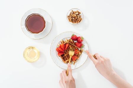 cropped view of woman cutting waffle on plate near cup with tea and bowl on whiteの写真素材