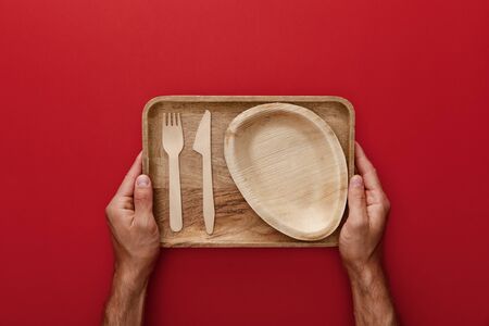 cropped view of man holding natural rectangular wooden dish with plate, fork and knife on red backgroundの写真素材